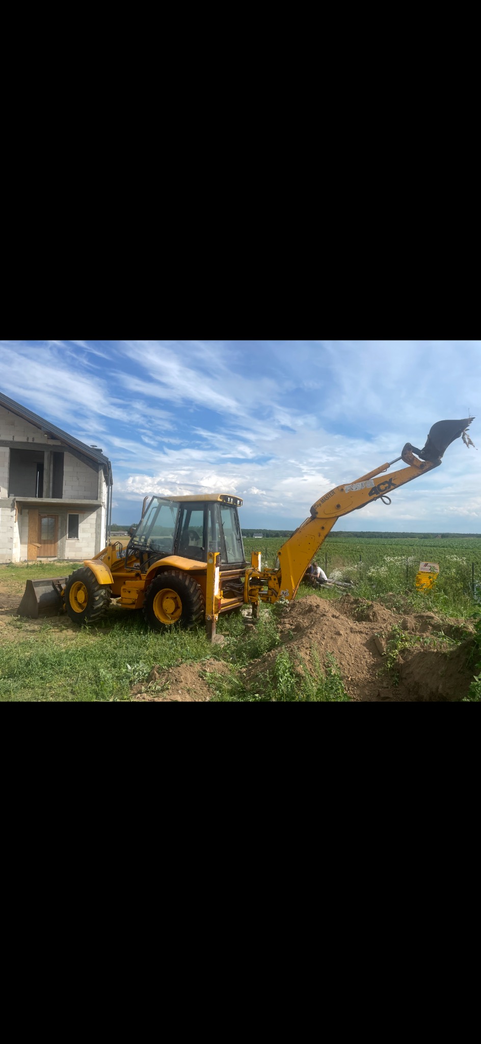 Żółta koparko-ładowarka JCB 4CX na trawiastym terenie, obok budynku w stanie surowym otwartym, wykonująca wykop. Błękitne niebo z chmurami w tle.