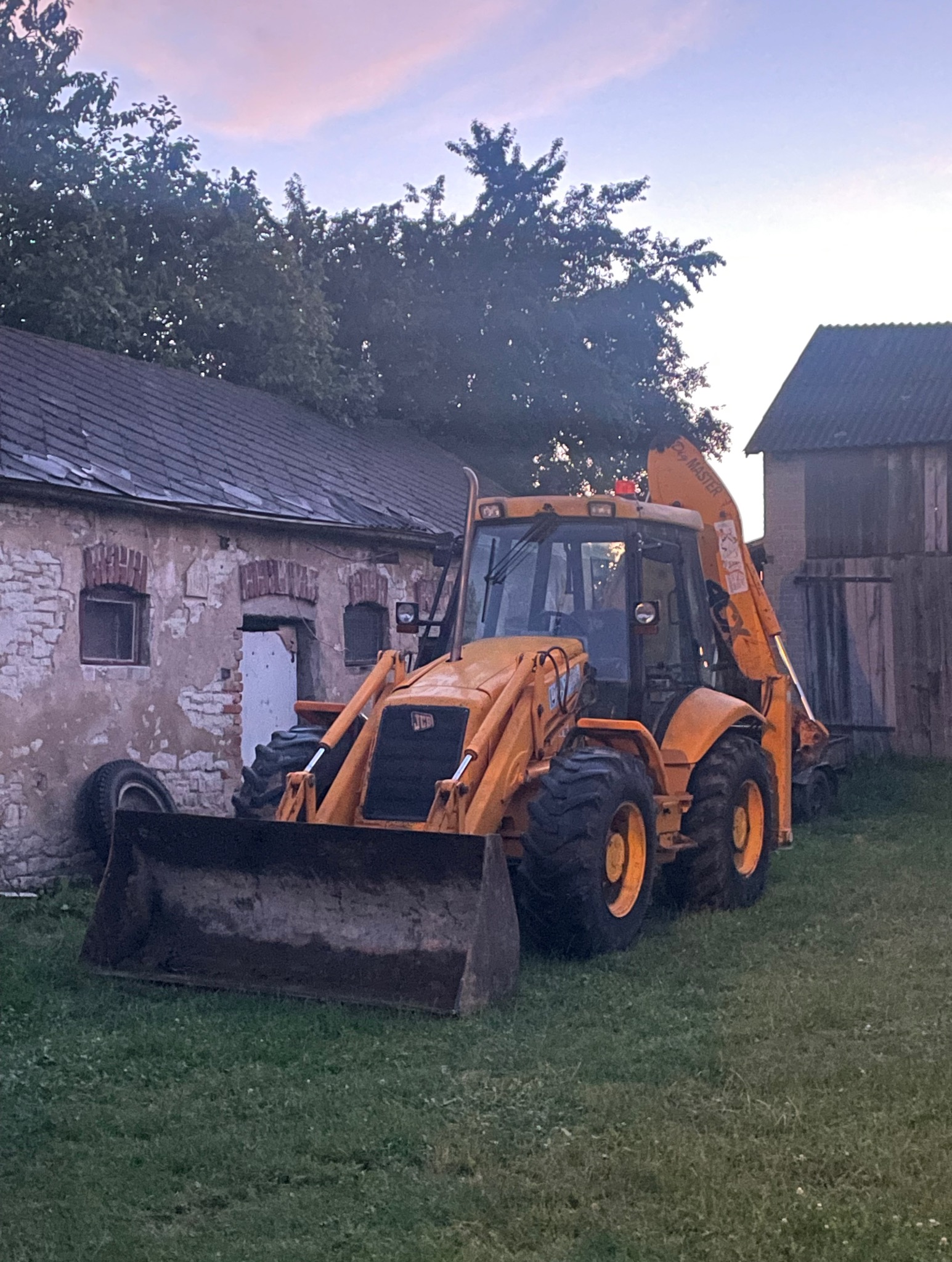 Żółta koparko-ładowarka JCB na trawie przed budynkiem z cegły i drewnianą stodołą w tle, uchwycona o zmierzchu.