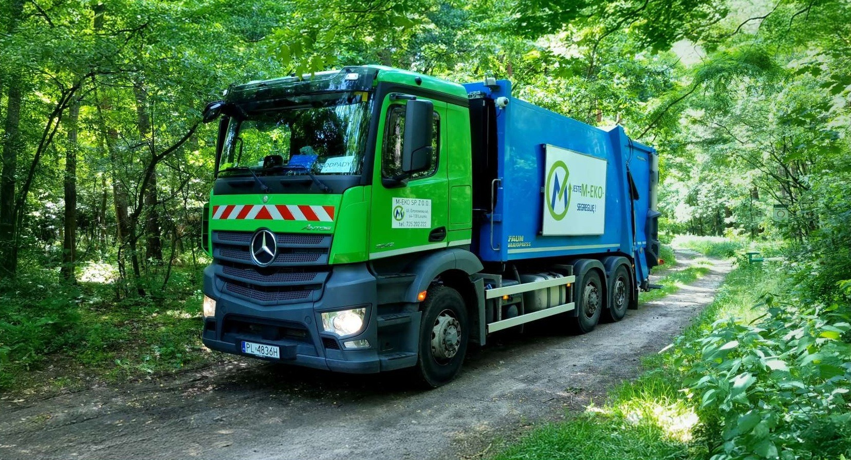 Zielono-niebieska śmieciarka z logo M-EKO na leśnej drodze, oświetlona promieniami słońca przebijającymi przez korony drzew. Selektywna zbiórka odpadów.