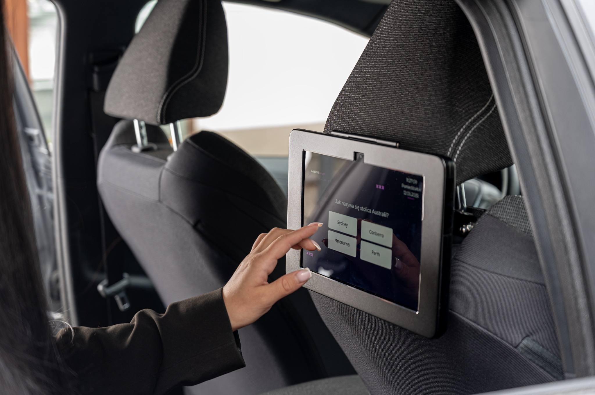 Ekran z quizem interaktywnym zamontowany na zagłówku fotela samochodowego, dotykany przez dłoń kobiety w czarnej marynarce. Widok z wnętrza pojazdu.