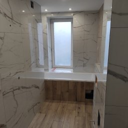 PHU Patryk Chwalisz - Wykończona łazienka w jasnych barwach z płytkami imitującymi marmur na ścianach i drewnopodobną podłogą. Widoczna wanna obudowana drewnem oraz okno.