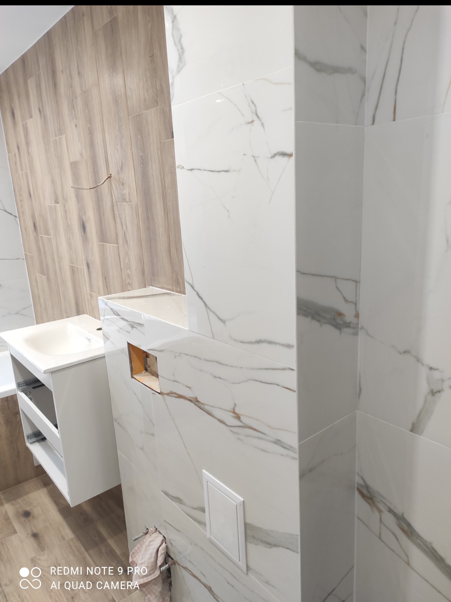 Biała umywalka z szafką w nowoczesnej łazience z marmurowymi płytkami i drewnopodobną ścianą. Widoczne elementy instalacji.