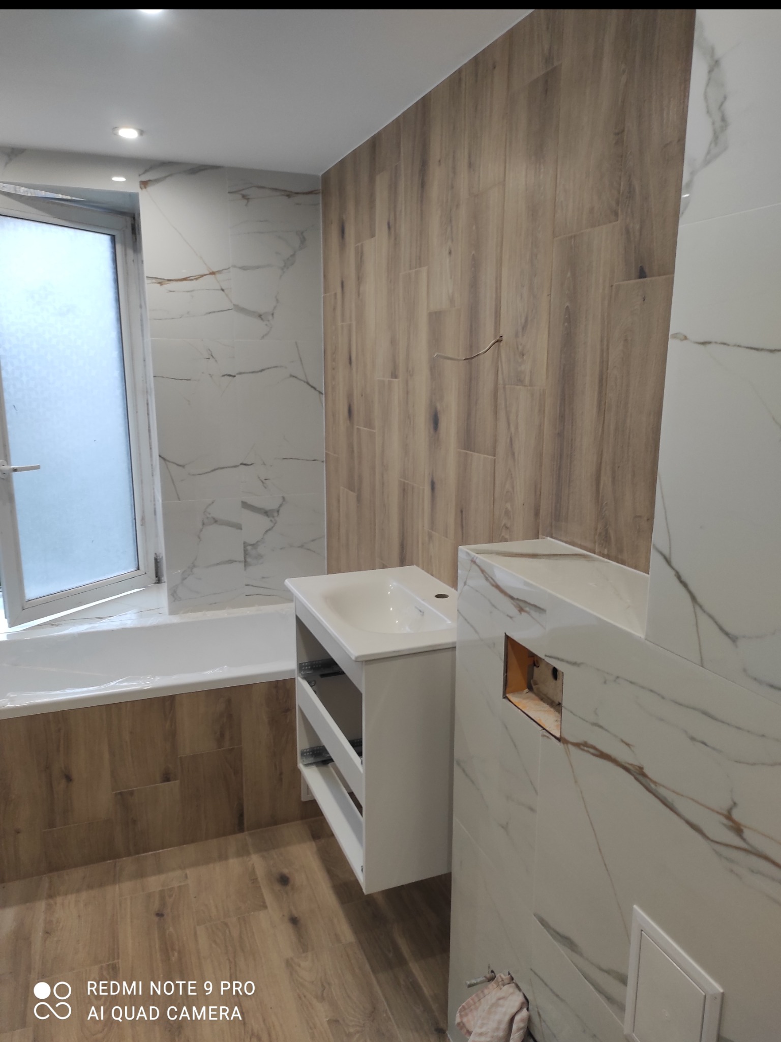 Nowoczesna łazienka po remoncie: białe marmurowe płytki, drewnopodobne panele ścienne i podłogowe, biała wanna i szafka pod umywalkę. Widoczne detale wykończenia.