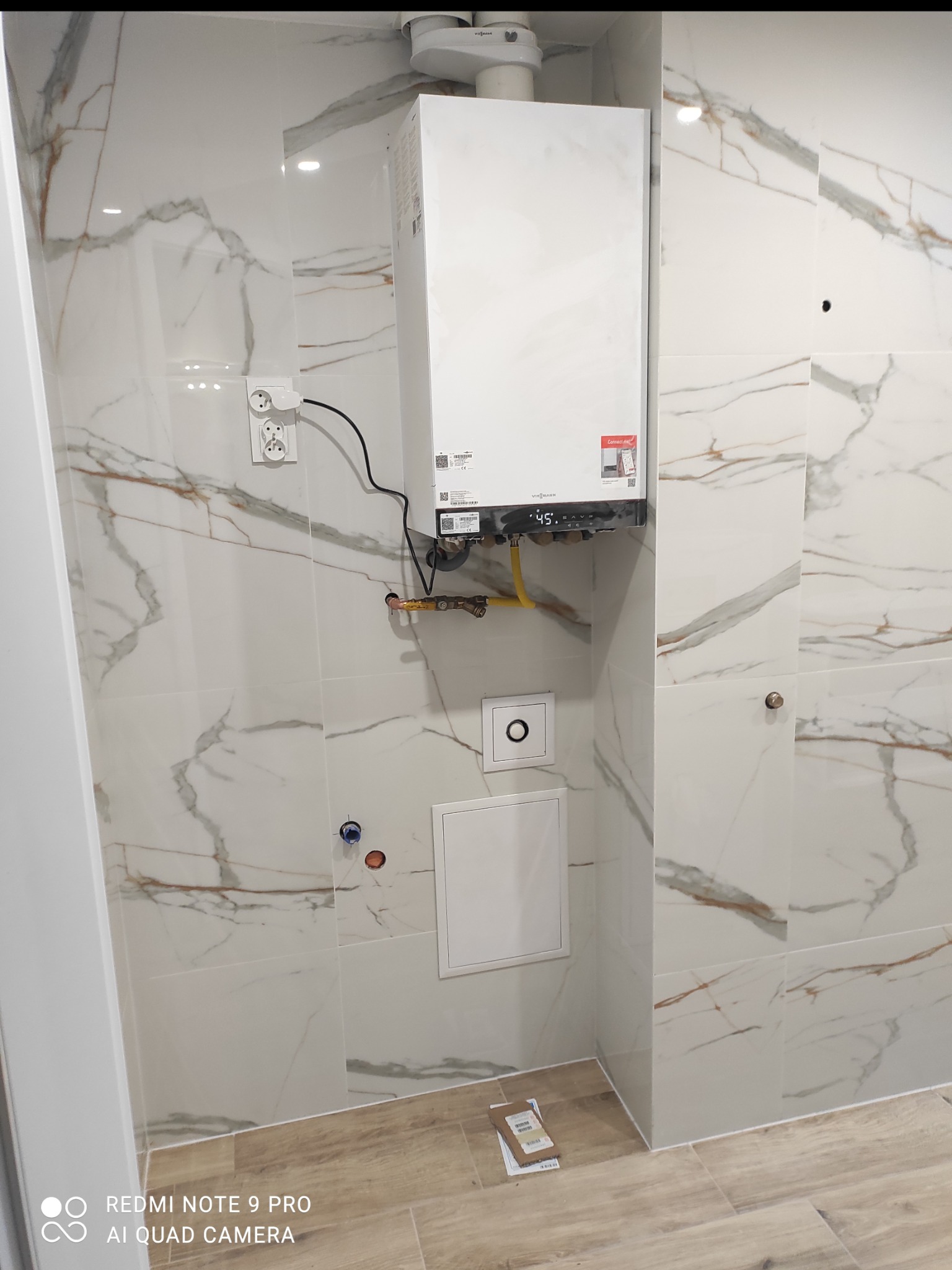 Biała łazienka z marmurowymi płytkami i zamontowanym piecem gazowym Viessmann. Widoczne gniazdka elektryczne i żółta rura gazowa.