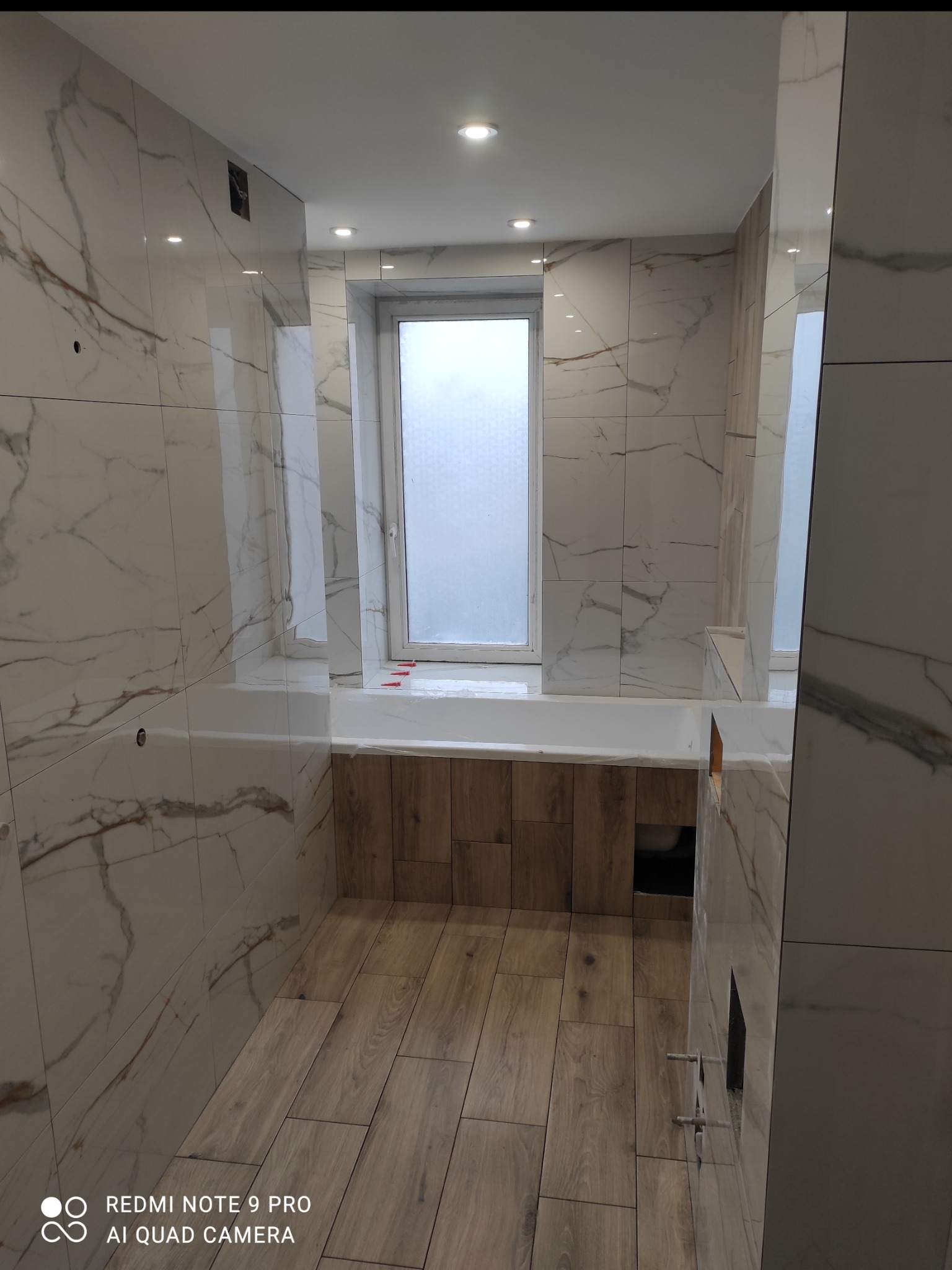 Wykończona łazienka w jasnych barwach z płytkami imitującymi marmur na ścianach i drewnopodobną podłogą. Widoczna wanna obudowana drewnem oraz okno.