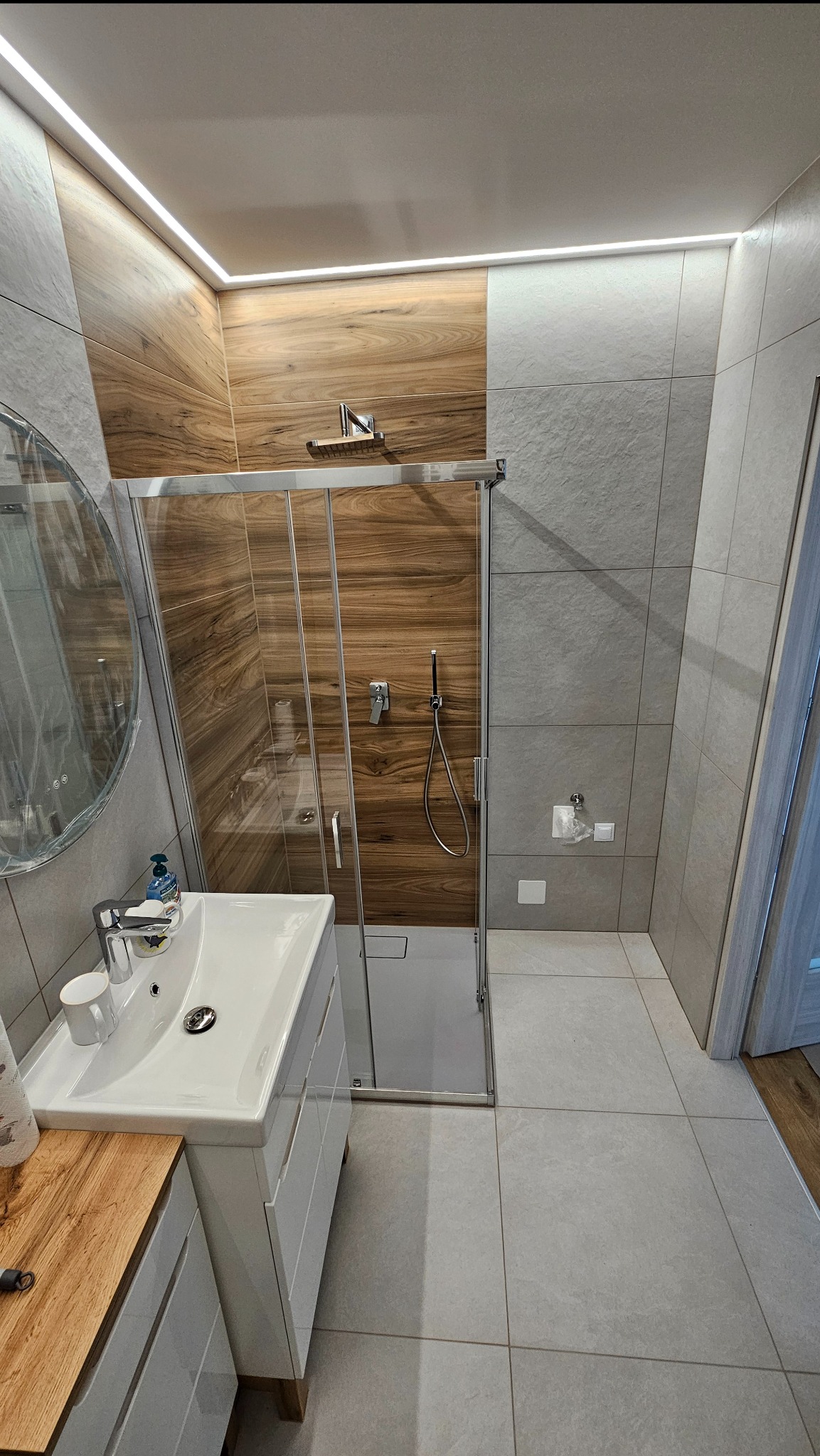 Nowoczesna łazienka z kabiną prysznicową, drewnopodobnymi i szarymi płytkami, oświetleniem LED podsufitowym i umywalką z szafką. Minimalistyczny design.