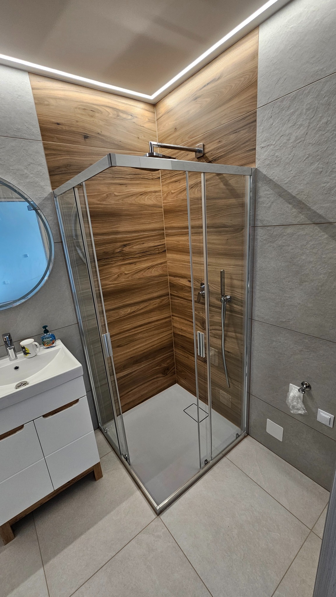 Nowoczesna kabina prysznicowa z drewnopodobnymi płytkami i subtelnym oświetleniem LED, umywalka z szafką w minimalistycznym stylu, szare płytki podłogowe.