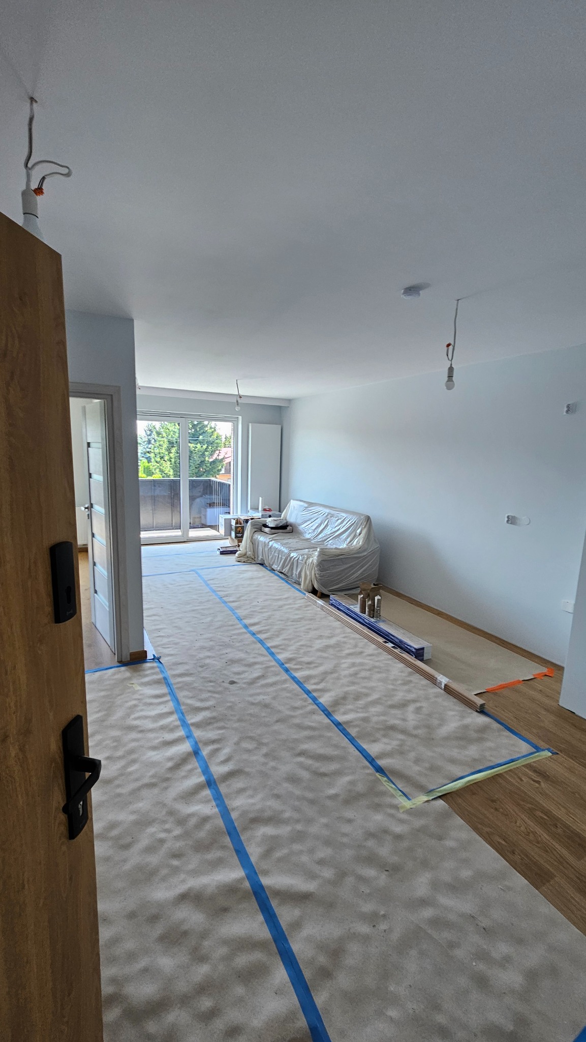 Jasne wnętrze w trakcie malowania: zabezpieczona podłoga, meble przykryte folią, świeżo pomalowane ściany, widoczne kable oświetleniowe. Przygotowanie do wykończenia.