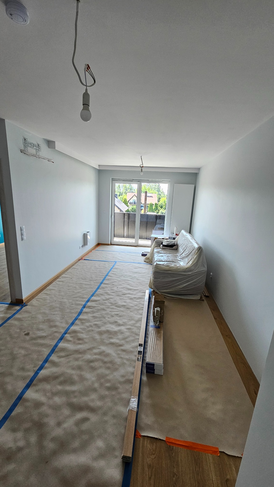 Pokój w trakcie remontu: zabezpieczona podłoga, ściany po malowaniu, okno balkonowe z widokiem na zieleń, sofa przykryta folią. Przygotowane panele podłogowe.