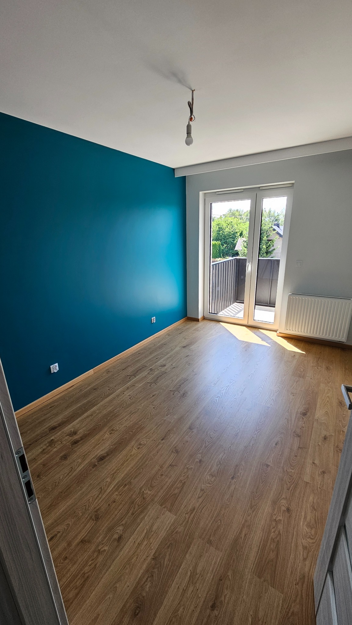 Pomalowane na turkusowo ściany i ułożone panele podłogowe w pokoju z balkonem. Widoczny grzejnik i niedokończone oświetlenie sufitowe.