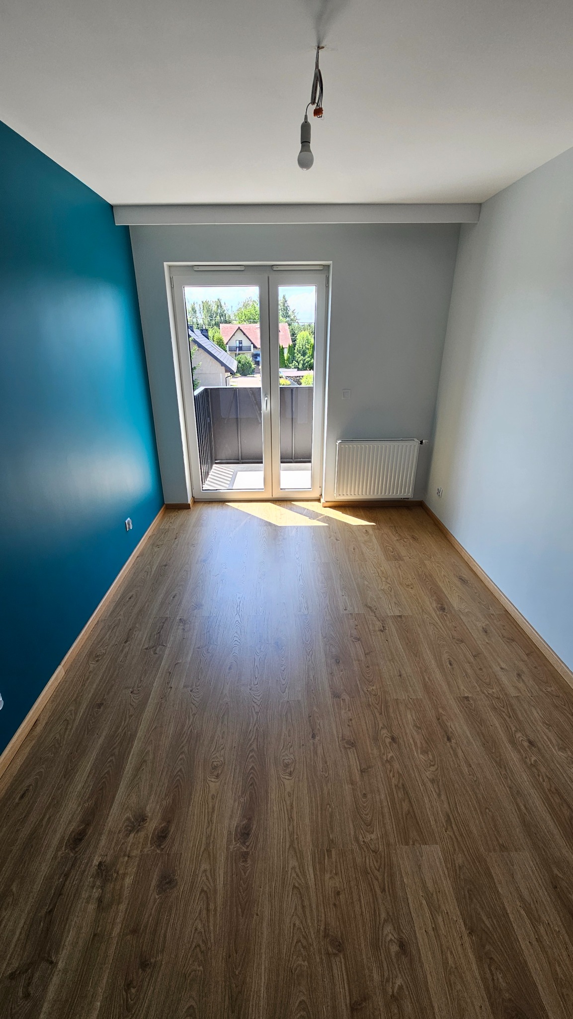 Pomalowane na turkusowo ściany, ułożone panele podłogowe w pustym pokoju z balkonem i wiszącą żarówką. Widok na zewnątrz przez otwarte drzwi balkonowe.