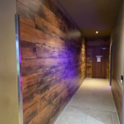 Korytarz z drewnianą ścianą, oświetlony fioletowym światłem. Widoczna podłoga z jasnych płytek i drzwi na końcu korytarza z napisem 'Herren'.