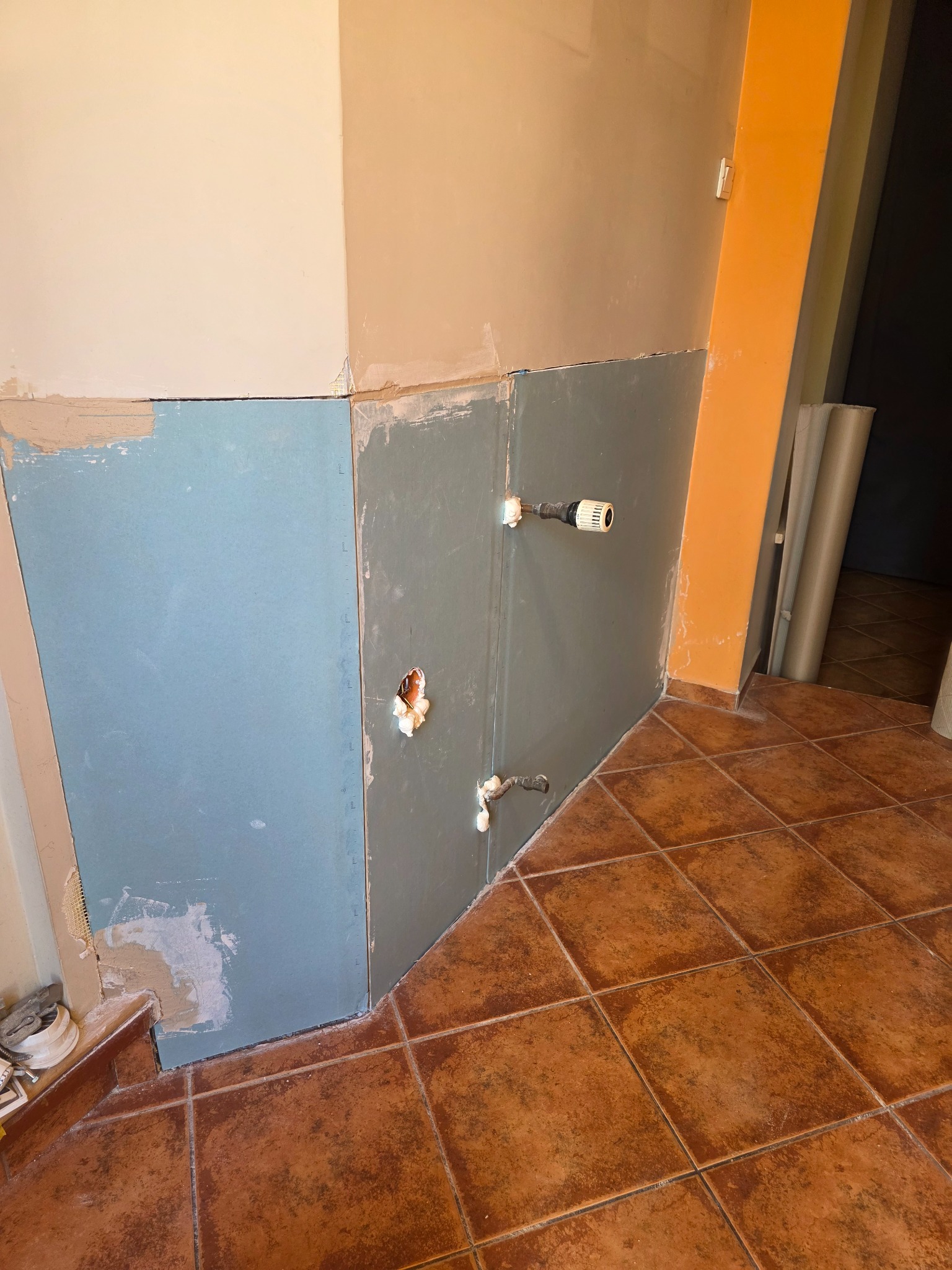 Płyty GK na ścianie w trakcie montażu, widoczne instalacje hydrauliczne i brązowe płytki podłogowe. Etap prac przygotowawczych w pomieszczeniu.