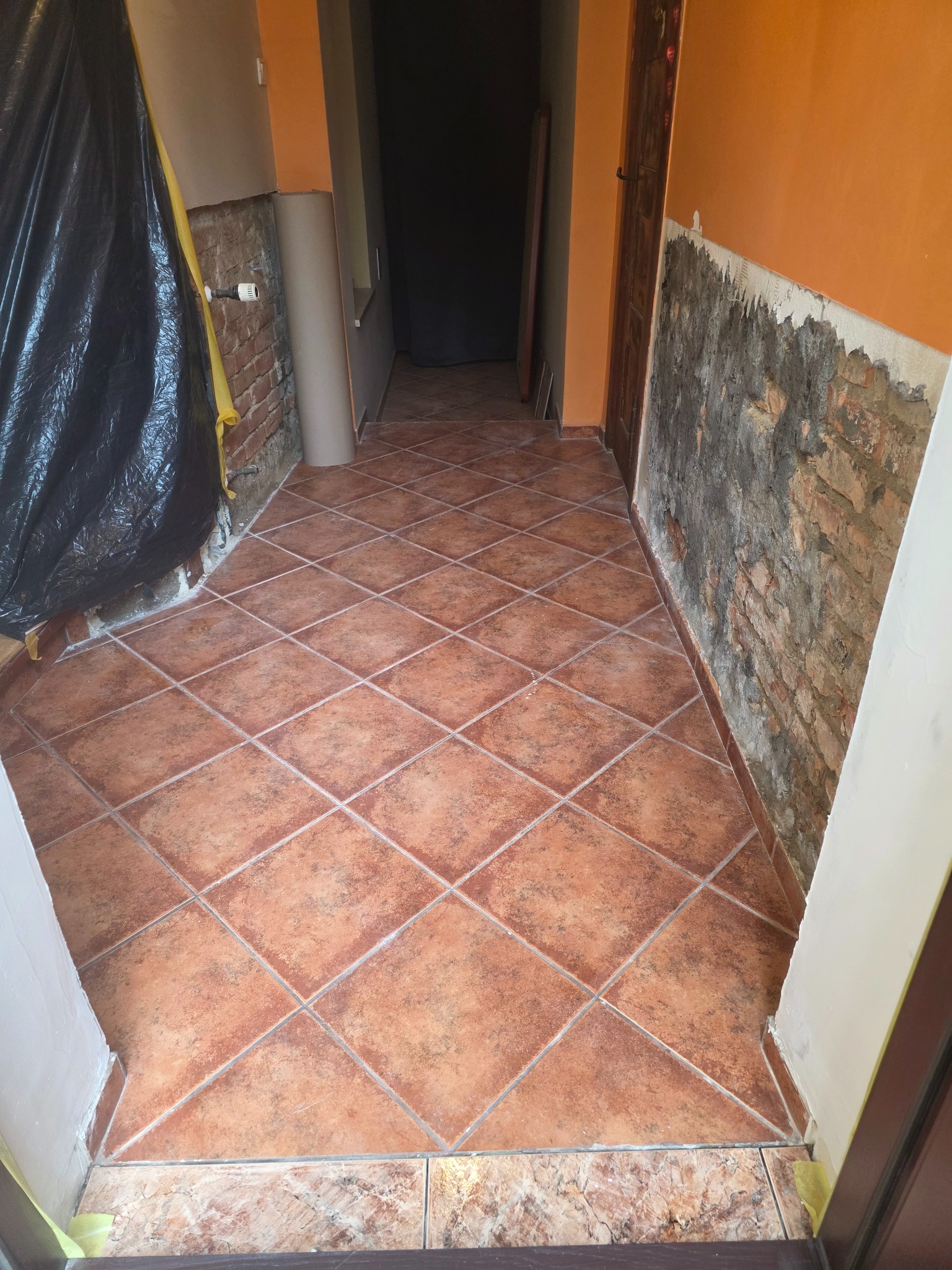Układanie terakoty w korytarzu podczas remontu. Widoczne przygotowane ściany do malowania i zabezpieczona folią część pomieszczenia. Kafelki ułożone w karo.