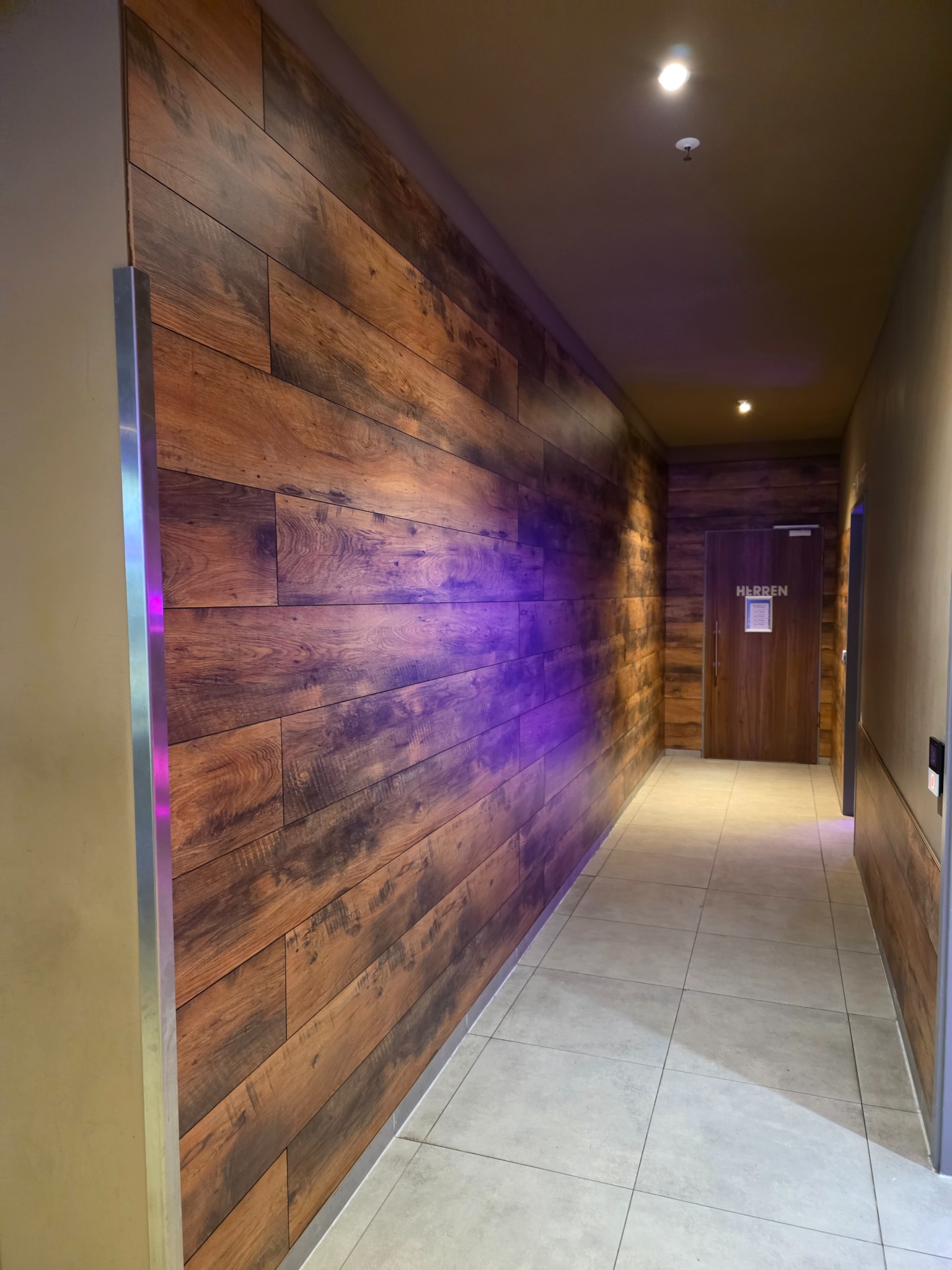 Korytarz z drewnianą ścianą, oświetlony fioletowym światłem. Widoczna podłoga z jasnych płytek i drzwi na końcu korytarza z napisem 'Herren'.