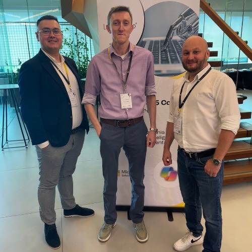 Trzech mężczyzn pozuje na tle roll-upu Microsoft Solutions z motywem laptopa i robota, prawdopodobnie podczas wydarzenia branżowego związanego z IT.