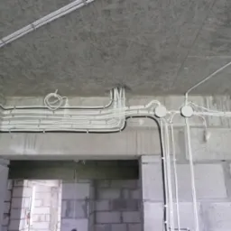 Białe i czarne przewody elektryczne ułożone na suficie i ścianach surowego wnętrza w trakcie budowy, widoczne puszki instalacyjne.