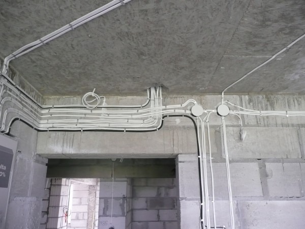 Białe i czarne przewody elektryczne ułożone na suficie i ścianach surowego wnętrza w trakcie budowy, widoczne puszki instalacyjne.