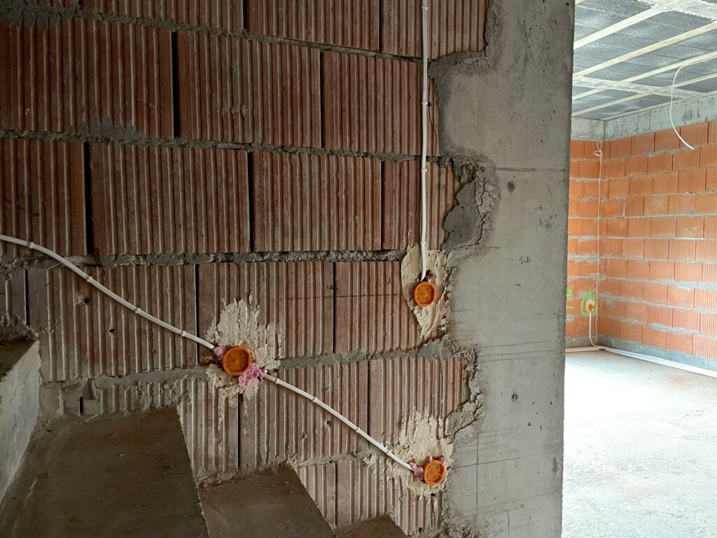Wewnętrzna instalacja elektryczna w surowym budynku: kable w peszlach, pomarańczowe puszki w ścianie z cegły kratówki. Widoczne fragmenty stropu i ścian.