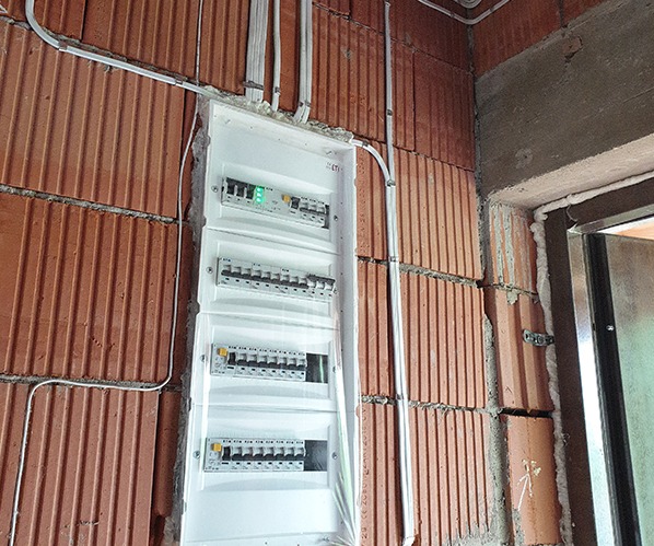 Rozdzielnica elektryczna w surowym murze z cegły, z widocznymi przewodami w białych rurkach. Widok na włączniki i bezpieczniki w białej obudowie.