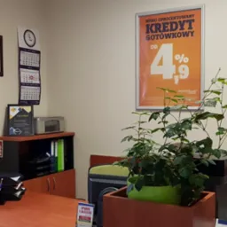 Wnętrze biura z plakatem reklamowym kredytu gotówkowego MerituBank, certyfikatem na ścianie, kalendarzem, biurkiem z dokumentami, kwiatem doniczkowym i nawilżaczem powietrza przy oknie.