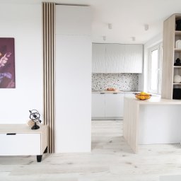 Magdalena Kozanko - Jasne, nowoczesne wnętrze: minimalistyczna kuchnia z wyspą, białe szafki z ryflowanym frontem, dekoracyjny panel z drewna i obraz z motywem roślinnym.