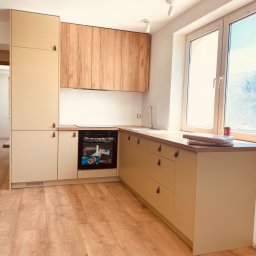 Magdalena Kozanko - Jasna kuchnia z drewnianymi akcentami: beżowe szafki dolne i wysoka, drewniane szafki górne, okno z widokiem na miasto i drewnopodobna podłoga.