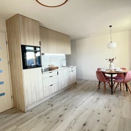 Magdalena Kozanko - Jasna kuchnia z drewnianymi frontami, okrągłym stołem i różowymi krzesłami. Białe drzwi z niebieskimi akcentami. Nowoczesne oświetlenie.