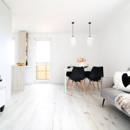 Magdalena Kozanko - Jasne, minimalistyczne wnętrze mieszkania z drewnianą podłogą, szarą sofą z poduszką w kształcie serca i jadalnią z czarnymi krzesłami. Na stole wazon z kwiatami.