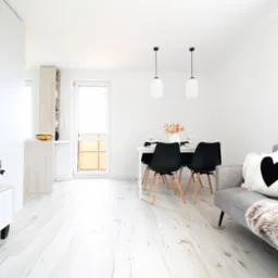 Jasne, minimalistyczne wnętrze mieszkania z drewnianą podłogą, szarą sofą z poduszką w kształcie serca i jadalnią z czarnymi krzesłami. Na stole wazon z kwiatami.