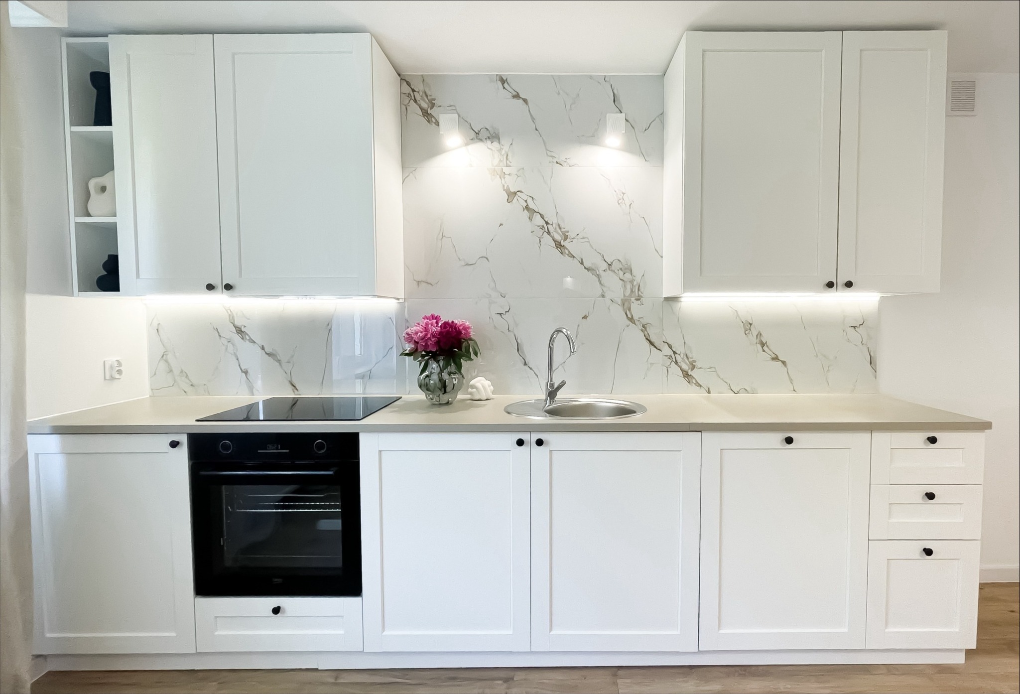 Biała, klasyczna kuchnia z marmurowym backsplashiem i kwiatami w wazonie. Ujęcie frontalne, prezentujące całą zabudowę kuchenną z piekarnikiem i płytą indukcyjną.