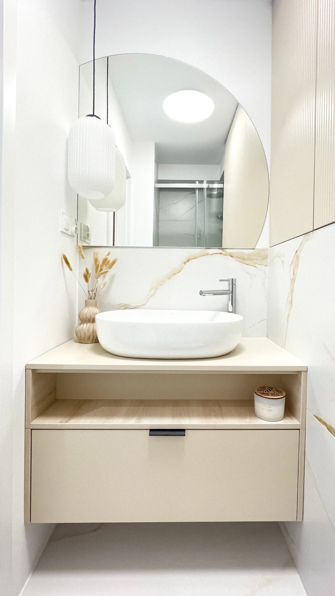 Nowoczesna łazienka z owalną umywalką nablatową, lustrem z łukiem i beżową szafką z szufladą. Widoczne akcenty dekoracyjne i fragment kabiny prysznicowej w odbiciu lustra.