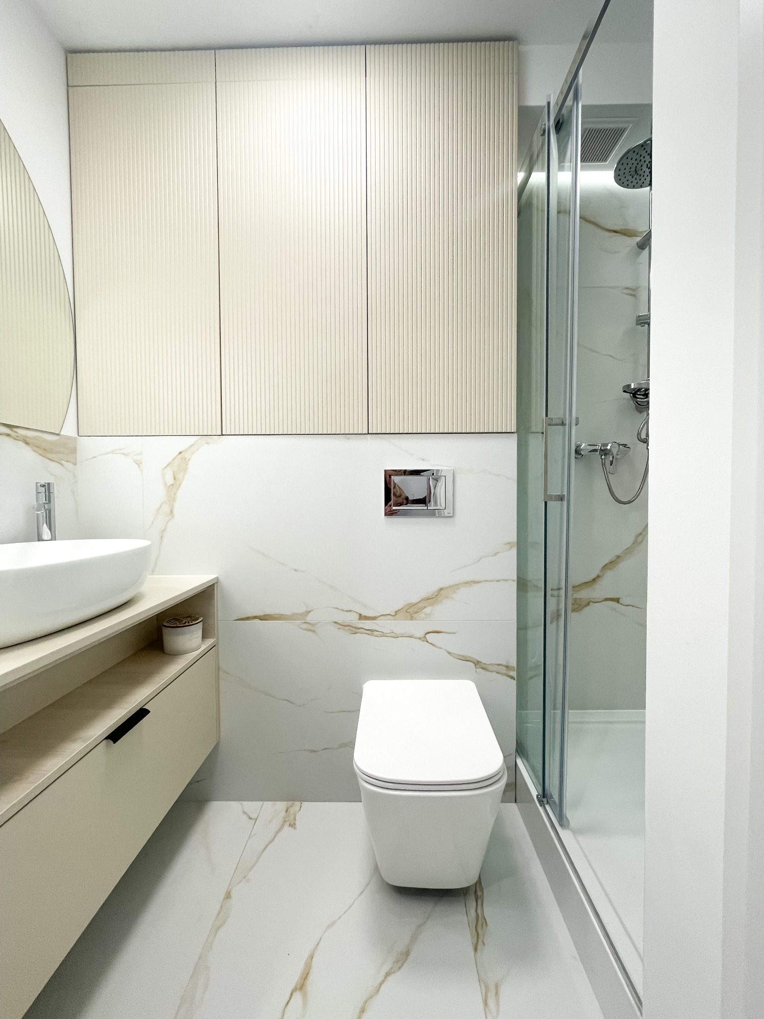 Nowoczesna łazienka w bieli i beżu: kabina prysznicowa, wisząca toaleta, umywalka nablatowa i szafki z ryflowanymi frontami na tle płytek imitujących marmur.