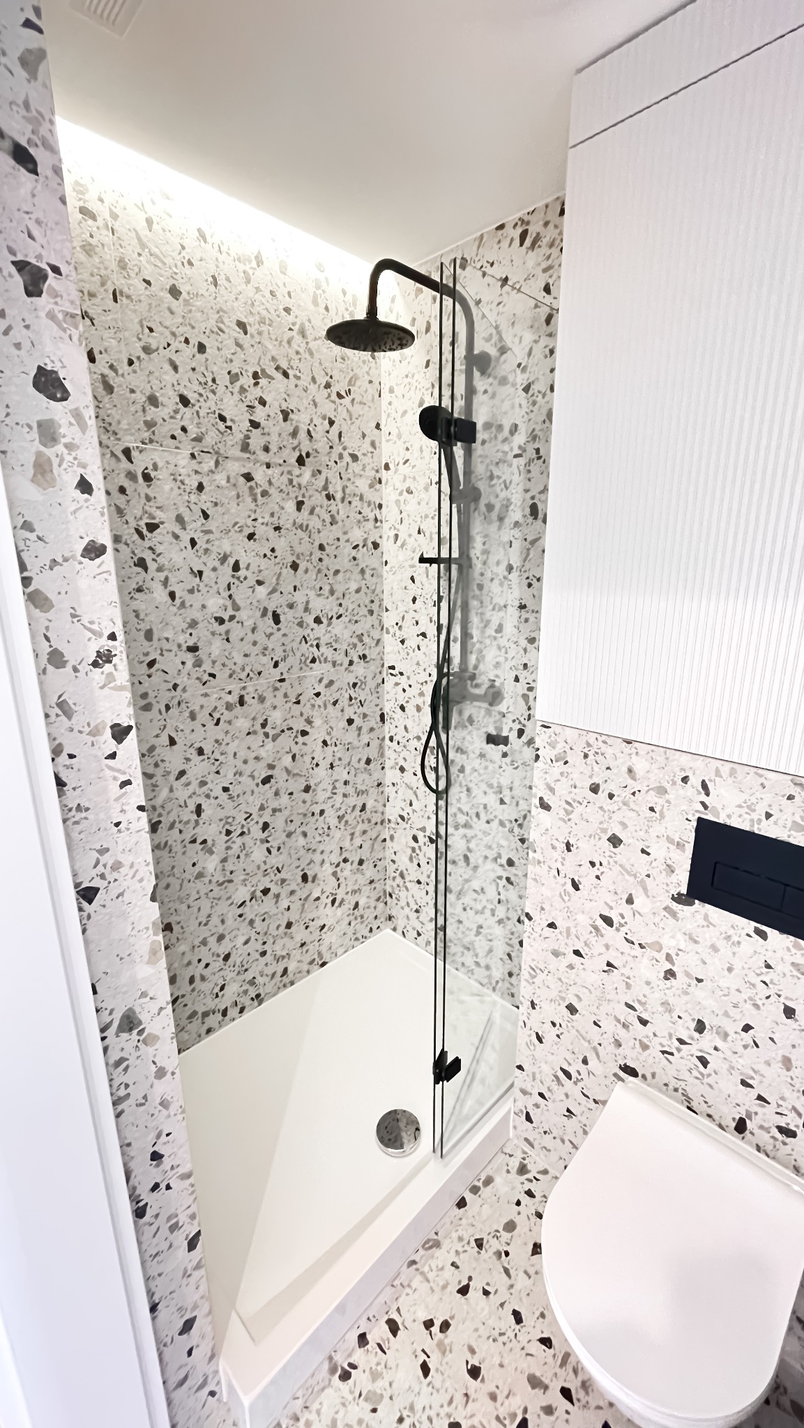 Nowoczesna kabina prysznicowa walk-in z czarną armaturą, biały brodzik i toaleta w łazience wykończonej płytkami lastryko. Minimalistyczny design.