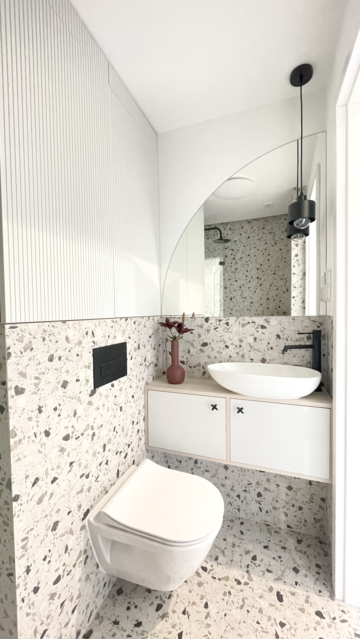 Nowoczesna łazienka z umywalką nablatową, wiszącą toaletą i płytkami lastryko na ścianach i podłodze. Lustro z zaokrąglonym łukiem. Minimalistyczne oświetlenie.