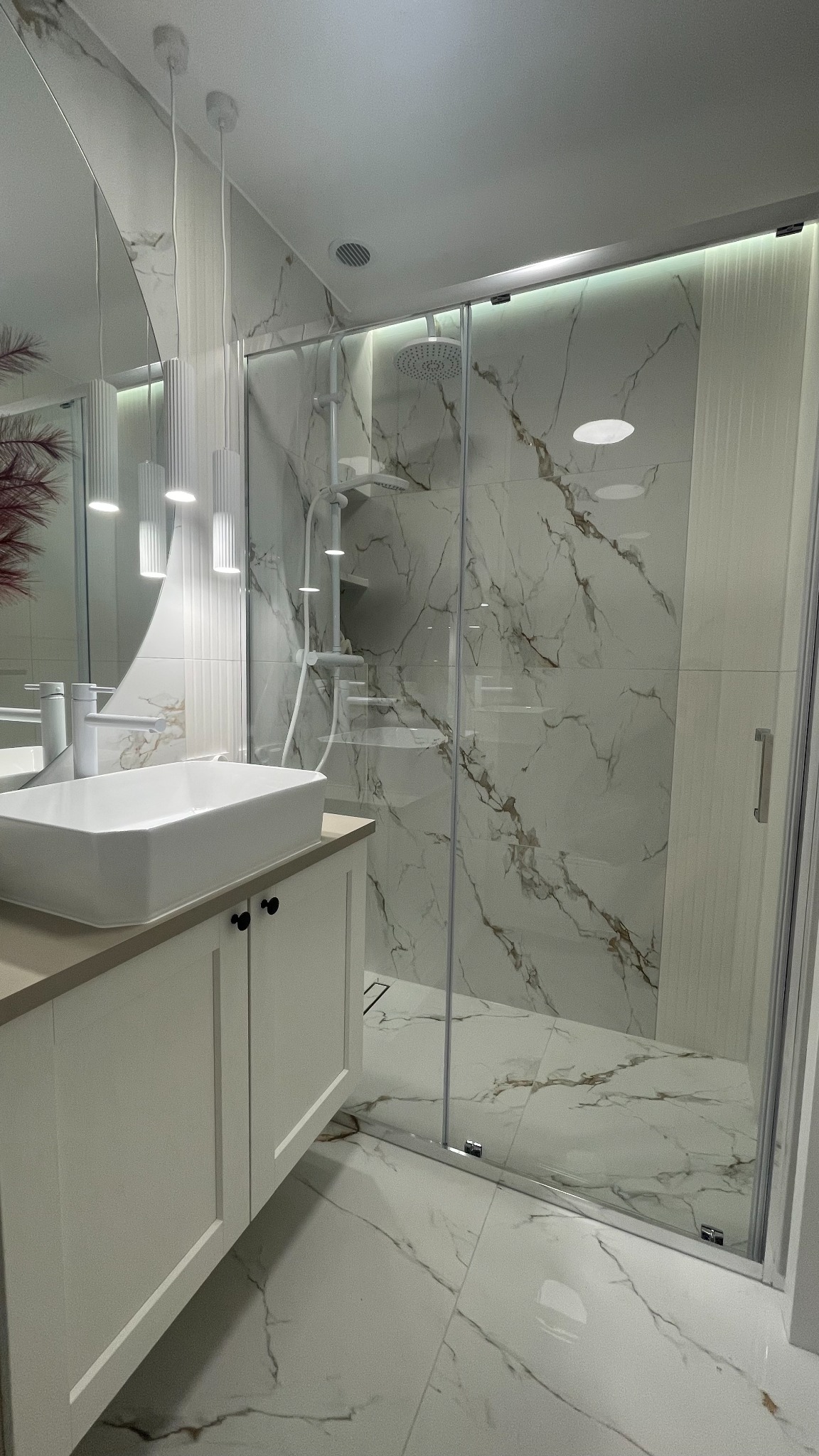 Nowoczesna łazienka z kabiną prysznicową typu walk-in, umywalką nablatową na szafce i lustrem. Białe płytki imitujące marmur na ścianach i podłodze.