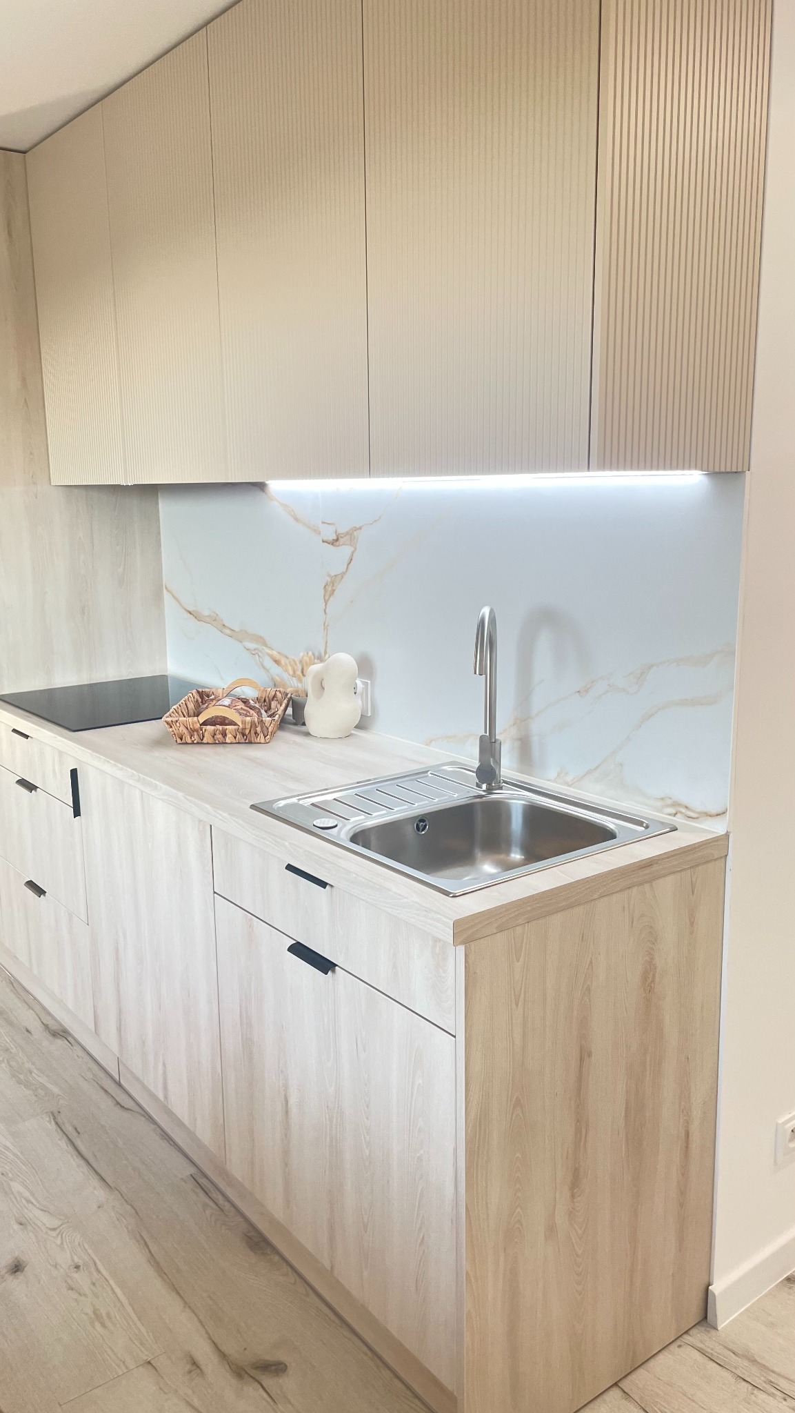 Jasna, nowoczesna kuchnia z drewnopodobnymi frontami, marmurowym panelem i oświetleniem LED. Ujęcie na zlew i blat z dekoracjami.