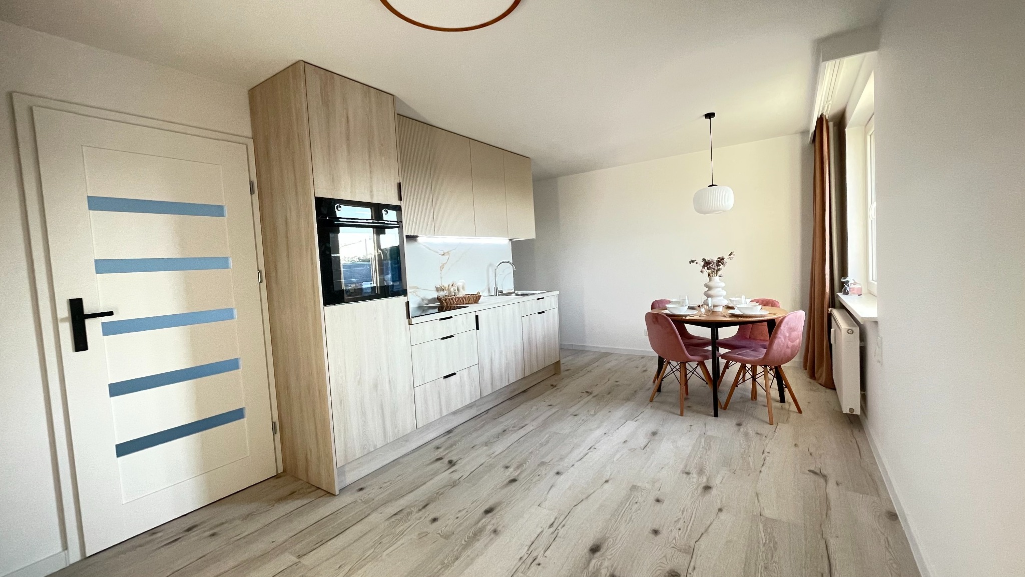 Jasna kuchnia z drewnianymi frontami, okrągłym stołem i różowymi krzesłami. Białe drzwi z niebieskimi akcentami. Nowoczesne oświetlenie.