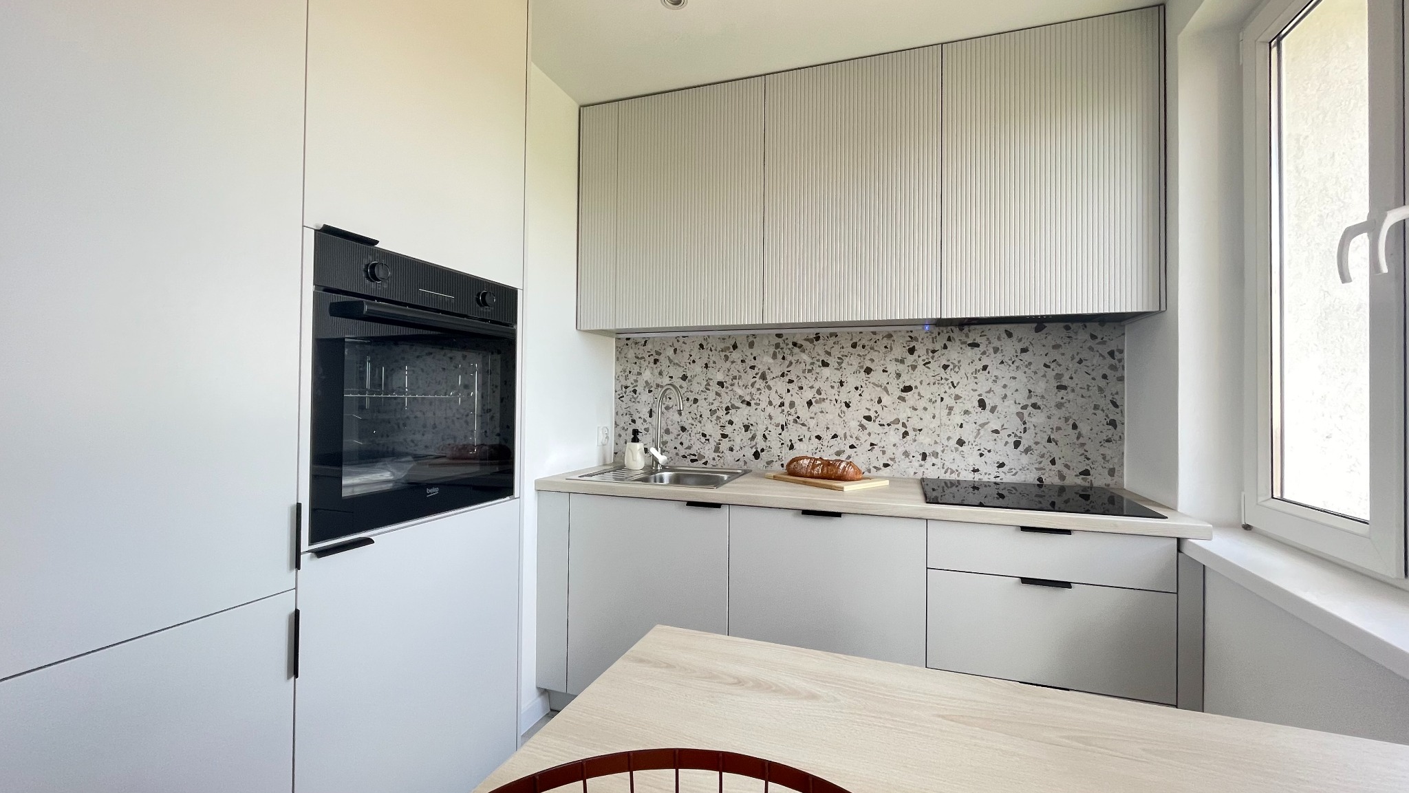 Jasna kuchnia z frezowanymi szafkami, mozaiką na ścianie i drewnianym blatem. Na blacie chleb na desce. Minimalistyczny design, okno z białymi ramami.