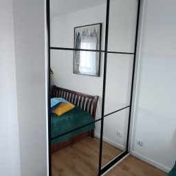 Tomaszewski Meble - Biała szafa wnękowa z przesuwnymi drzwiami lustrzanymi i czarnymi ramami, odbijająca wnętrze sypialni z drewnianym łóżkiem i obrazem na ścianie.