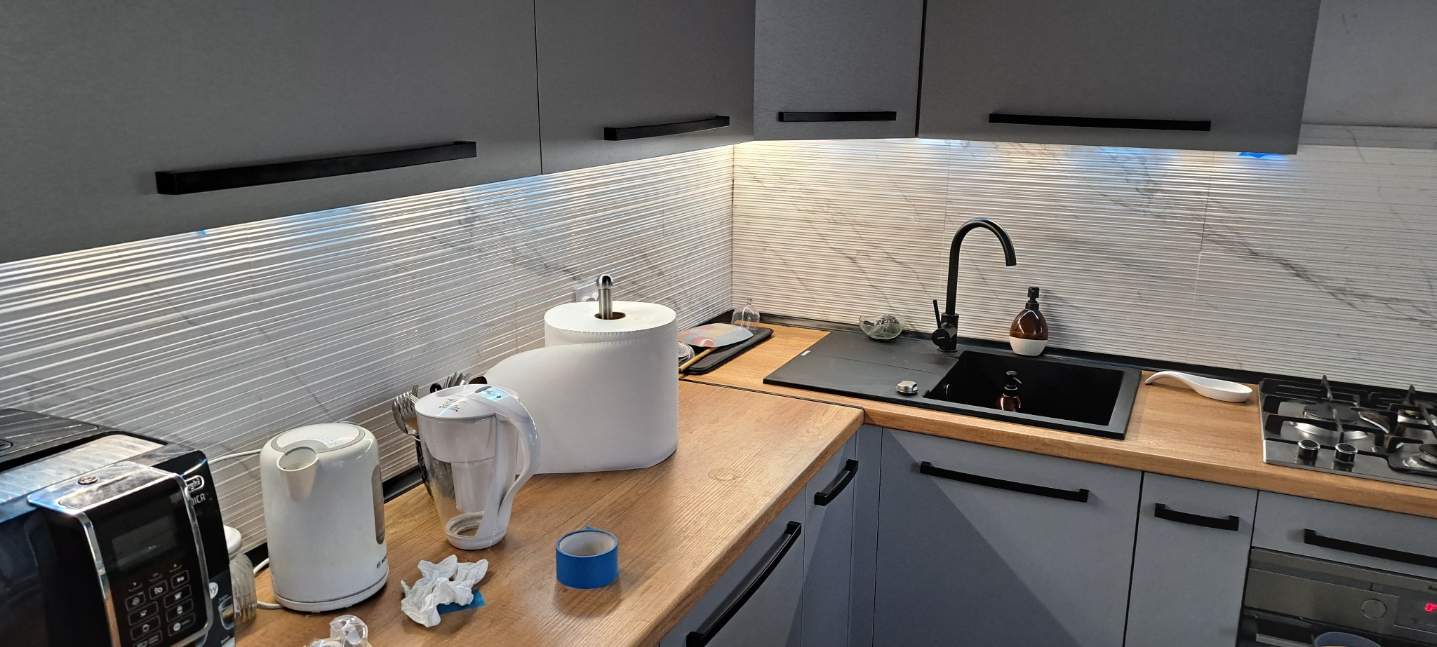 Szare szafki kuchenne z czarnymi uchwytami, drewniany blat, biała mozaika na ścianie i czarny zlew. Widoczny fragment ekspresu do kawy i akcesoria kuchenne.