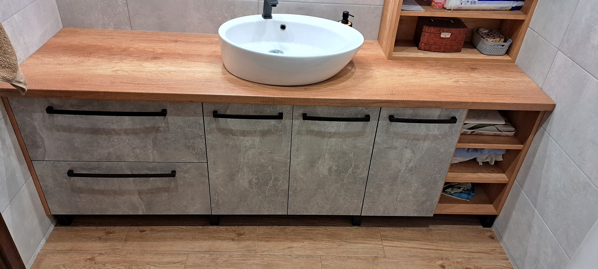 Nowoczesna szafka łazienkowa z blatem imitującym drewno, szarymi frontami i czarnymi uchwytami. Umywalka nablatowa, otwarte półki z tekstyliami.