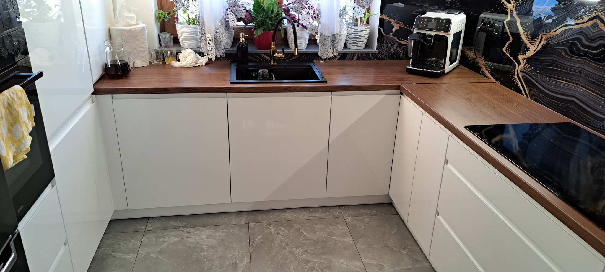 Nowoczesna kuchnia z białymi, lakierowanymi frontami i drewnianym blatem. Widoczny czarny zlew i złota bateria. Na blacie ekspres do kawy i płyta indukcyjna.