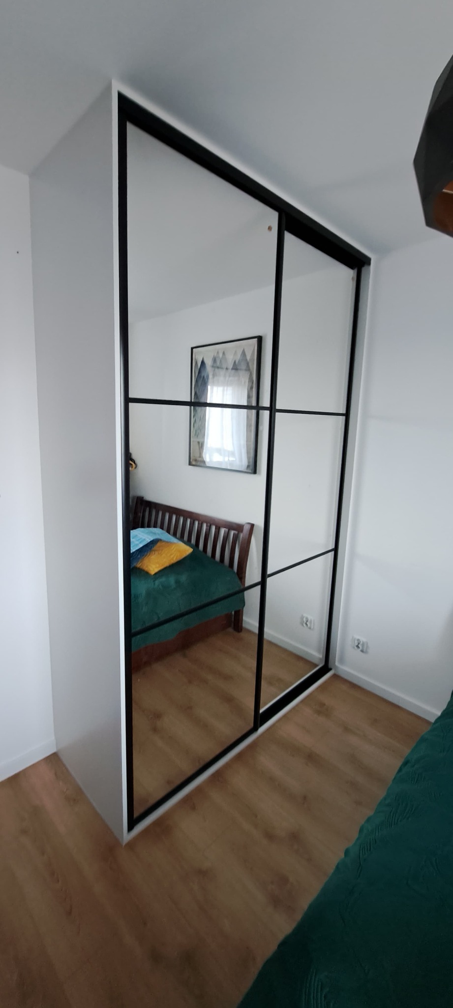 Biała szafa wnękowa z przesuwnymi drzwiami lustrzanymi i czarnymi ramami, odbijająca wnętrze sypialni z drewnianym łóżkiem i obrazem na ścianie.