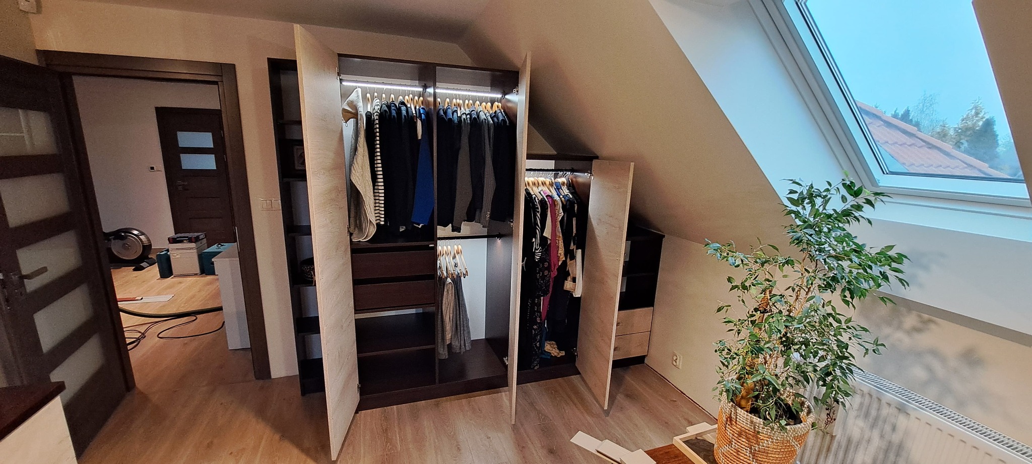Szafa wnękowa na poddaszu z oświetleniem LED, otwarte drzwi ukazują ubrania na wieszakach. Widoczny fragment okna dachowego i roślina doniczkowa.