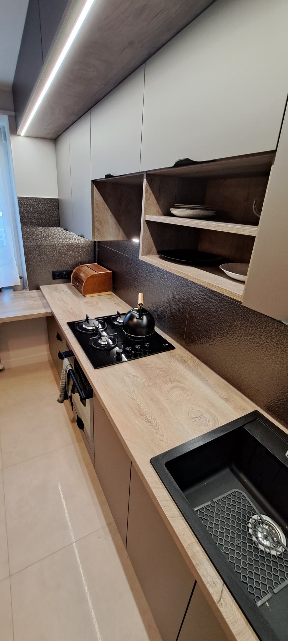 Nowoczesna kuchnia z drewnianym blatem, czarnym zlewem i płytą grzewczą. Na blacie czajnik, a nad nim półki z naczyniami. Uwagę przyciąga teksturowana ściana.