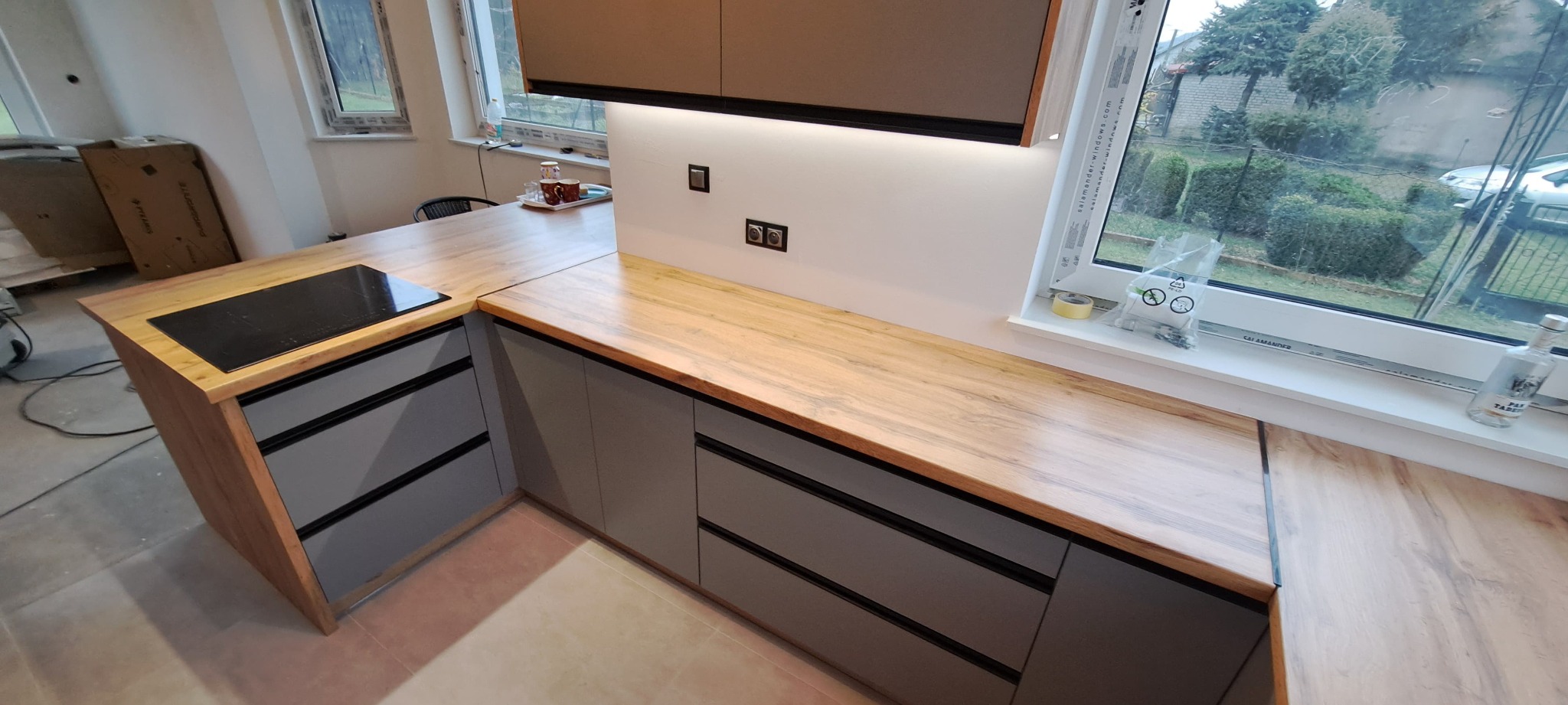 Nowoczesna kuchnia z drewnianym blatem i szarymi szafkami, płyta indukcyjna, okno z widokiem na ogród. Oświetlenie LED pod szafkami.