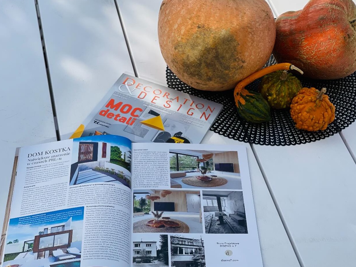 Magazyn wnętrzarski z aranżacjami domów i mieszkań, dekoracje z dyń na białym stole. Widoczne projekty domów w stylu PRL i nowoczesne wnętrza.