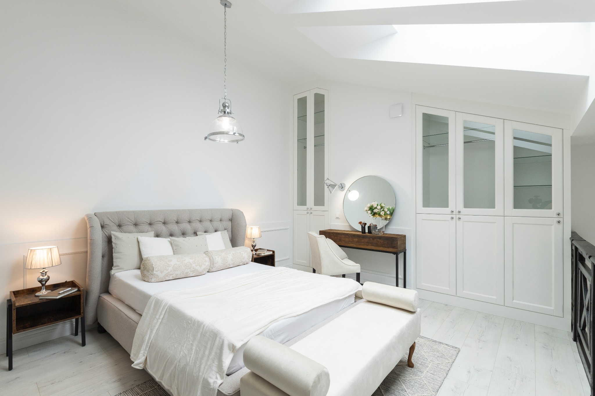 Jasna sypialnia z dużym łóżkiem, toaletką z okrągłym lustrem i wbudowaną szafą. Skosy i okno dachowe doświetlają wnętrze. Minimalistyczny design.
