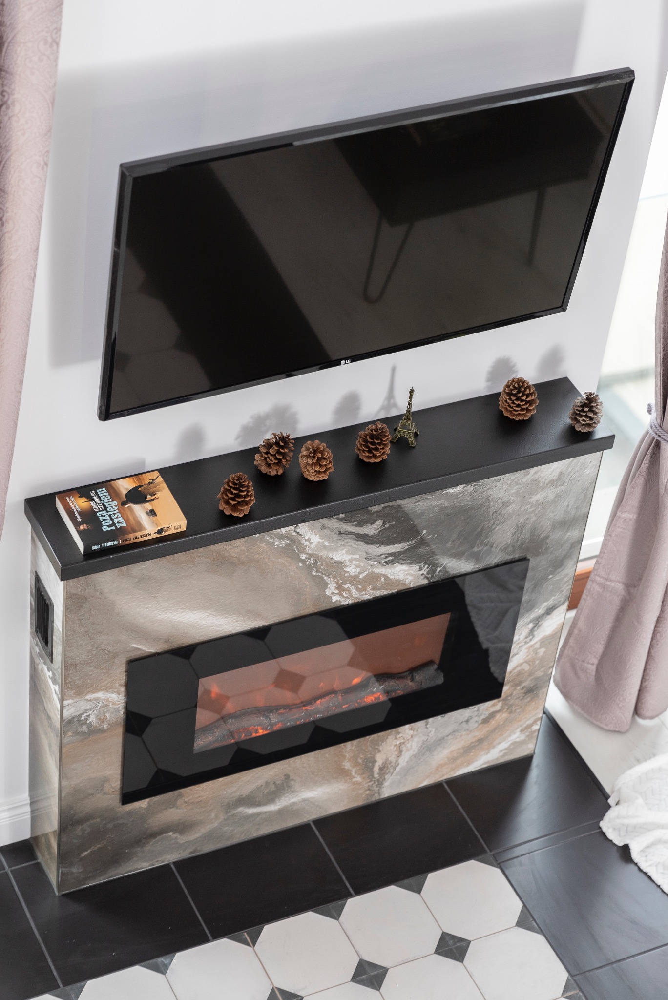 Nowoczesny kominek elektryczny z marmurowym wykończeniem, telewizorem LG nad nim, książką i szyszkami na blacie. Podłoga w biało-czarną szachownicę.